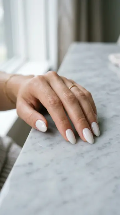 A close up beauty photograph of a woman's hand with short almond shaped nails painted in a soft, opaque milky white shade with a high gloss finish