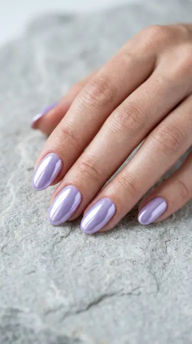 A close up photograph of a woman's hand with short almond shaped nails featuring a mirror like pastel chrome finish in soft lavender